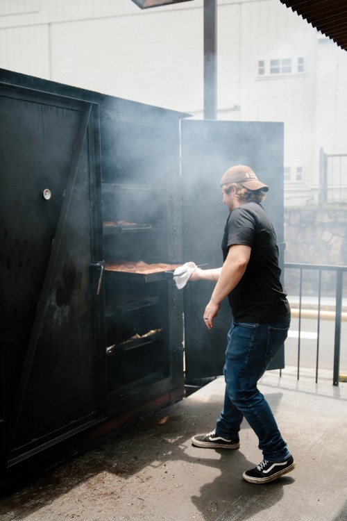 A person in a black shirt and cap removes food from a smoky outdoor smoker.