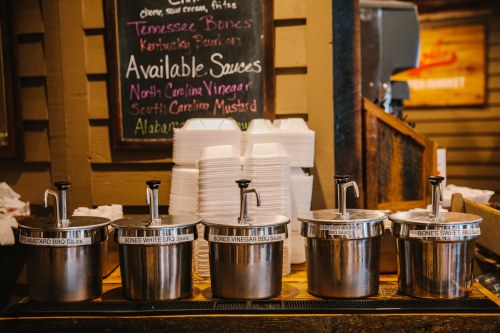 A wooden counter with four sauce dispensers labeled for BBQ sauces, alongside stacks of takeout containers.