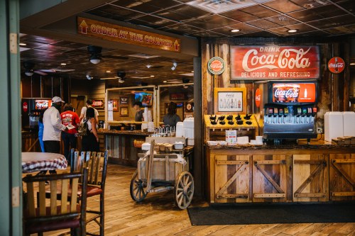 A rustic restaurant interior with a Coca-Cola sign, drink station, and customers ordering at the counter.