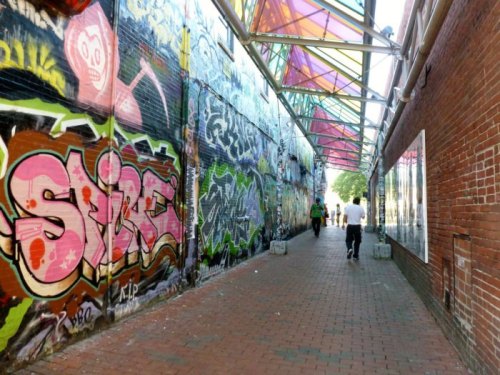 A vibrant alleyway adorned with colorful graffiti, featuring a glass canopy and people walking through.