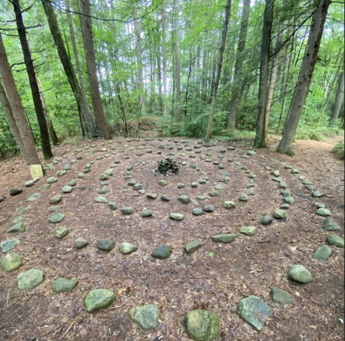 A stone labyrinth surrounded by trees in a forest, with a small pile of stones at the center.