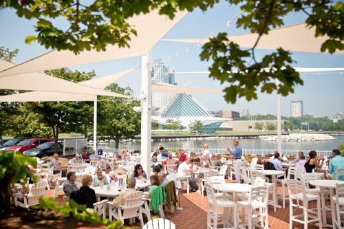 A lively outdoor dining area by the water, with people enjoying meals under shaded canopies and trees.
