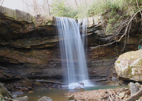 A serene waterfall cascades over rocky cliffs into a tranquil pool, surrounded by lush greenery and natural stone formations.