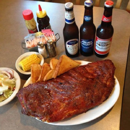 A large plate of BBQ ribs with sides of corn, salad, and potato wedges, accompanied by three bottles of beer.
