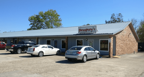 A brick building with a metal roof, featuring a sign for "Dempsey's" and parked cars in front on a sunny day.