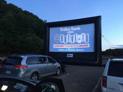 Inflatable movie screen at a drive-in event, with cars parked nearby under a twilight sky.