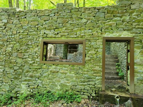 A stone wall with a window and doorway, leading to a staircase in a forested area with green foliage.