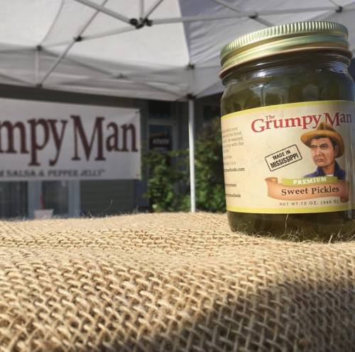 A jar of Grumpy Man sweet pickles on a burlap surface, with a banner in the background displaying the brand name.