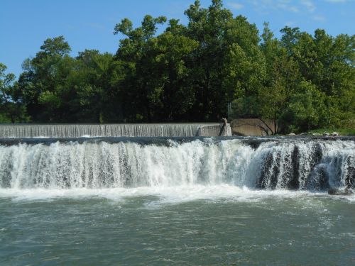 A serene waterfall cascading over a dam, surrounded by lush green trees under a clear blue sky.