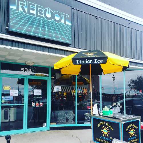 A storefront with a sign reading "REBOOT" and an outdoor cart selling Italian ice under a yellow and black umbrella.
