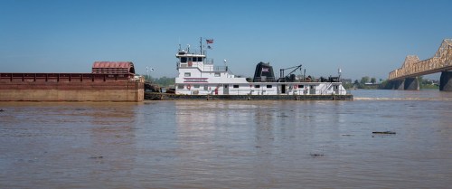 A towboat pushes a barge along a river, with a bridge visible in the background under a clear blue sky.