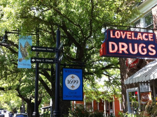 Street signs in a town, featuring directions to Shearwater Pottery and Mary C. O'Keefe, with a Lovelace Drugs sign nearby.