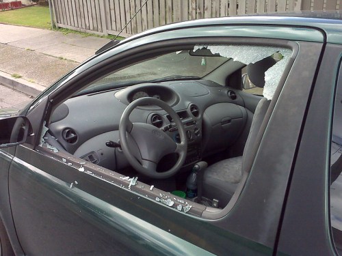 A car with a broken window, shattered glass on the seat and ground, indicating possible vandalism or theft.