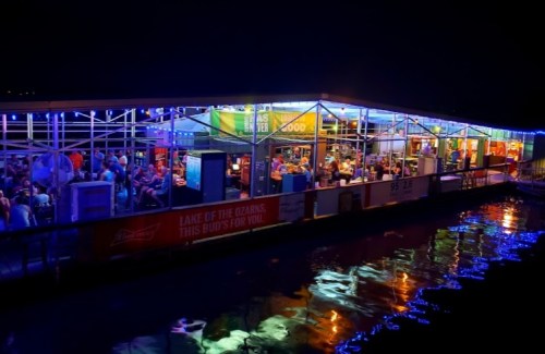 A lively lakeside restaurant at night, illuminated with colorful lights and filled with diners enjoying the atmosphere.