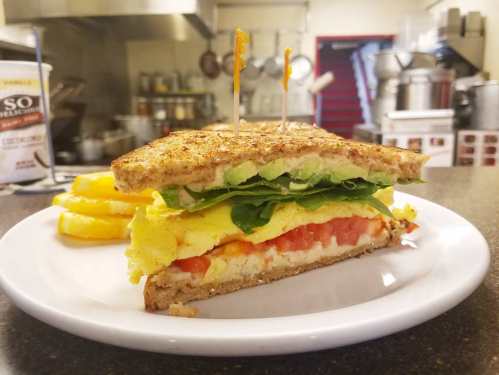 A hearty sandwich with layers of egg, spinach, tomato, and cucumber on toasted bread, served with pineapple slices.
