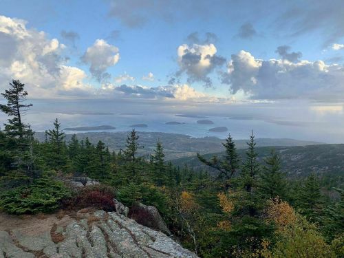 A scenic view from a mountain overlooking islands and a body of water, surrounded by trees and clouds in the sky.