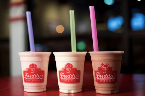 Three milkshakes in cups with Freddy's logo, featuring colorful straws, on a red table.