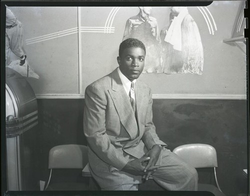 A man in a suit sits on a chair, with a vintage jukebox and wall art in the background.
