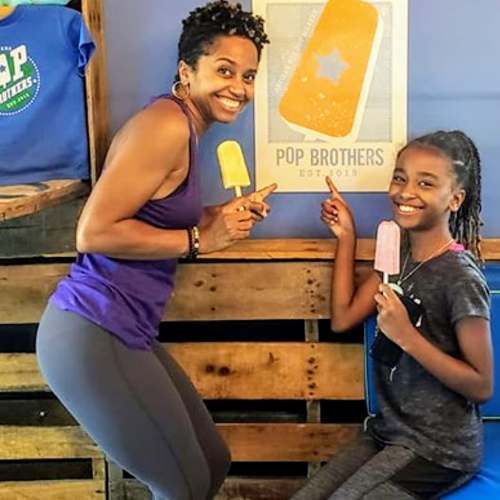 A woman and a girl smile and pose with colorful popsicles in front of a "Pop Brothers" sign.