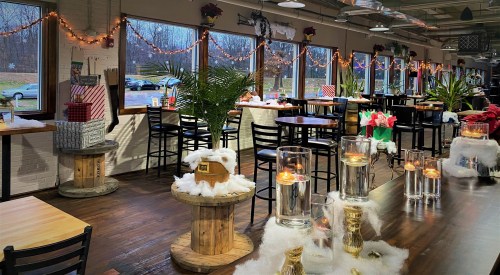 Cozy restaurant interior decorated for the holidays, featuring tables, candles, and festive decor with gifts and greenery.