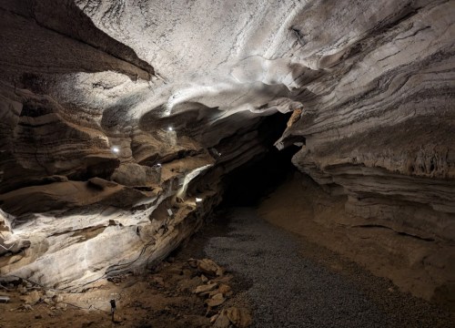 A dimly lit cave with textured rock formations and a gravel floor, leading into a dark passageway.