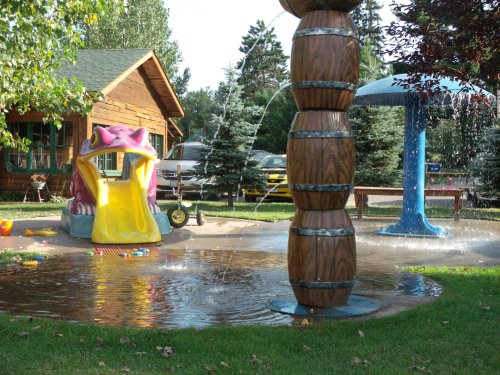 A colorful water play area featuring a frog slide, water fountains, and a wooden cabin in the background.