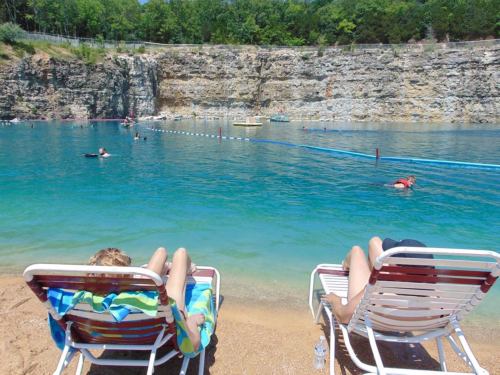 Two people relax on beach chairs by a clear blue lake, with swimmers enjoying the water and rocky cliffs in the background.