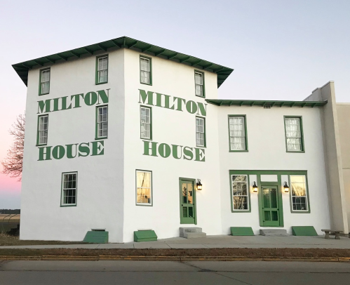 A two-story white building with green trim, featuring large text that reads "MILTON HOUSE" on its exterior walls.