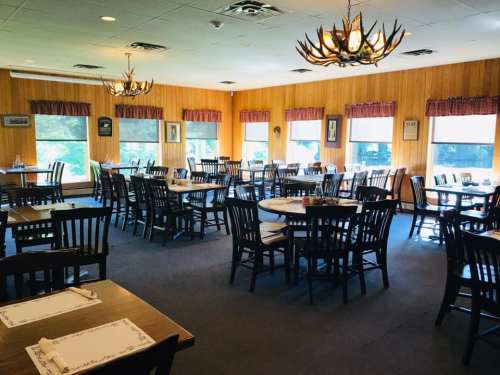 A spacious restaurant interior with wooden walls, multiple tables, and large windows letting in natural light.
