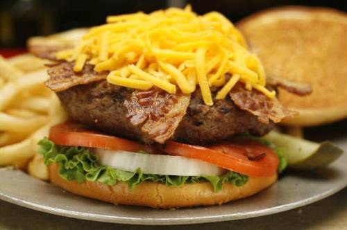 A close-up of a burger topped with crispy bacon and cheddar cheese, served with lettuce, tomato, and fries on the side.