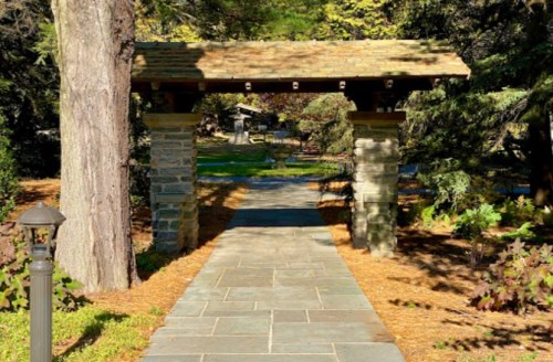 A stone pathway leads through a garden archway, surrounded by trees and greenery on a sunny day.