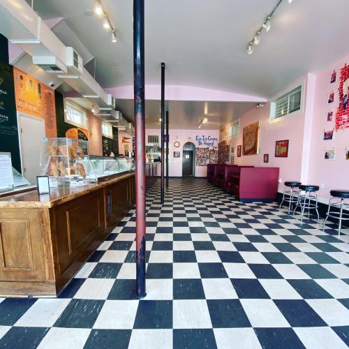 Interior of an ice cream shop with a checkered floor, wooden counter, and pink walls adorned with artwork.