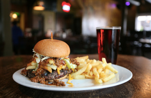 A hearty burger topped with chili, cheese, and lettuce, served with fries and a glass of dark beer on a wooden table.