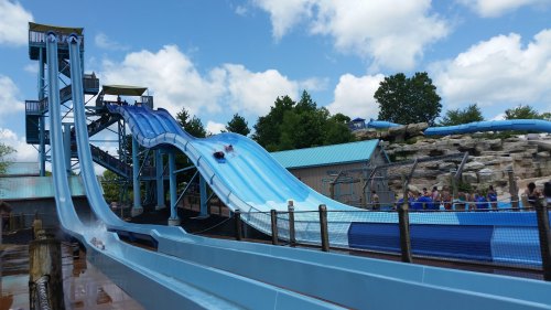 Two water slides at a water park, with people sliding down and a crowd enjoying the area under a blue sky.