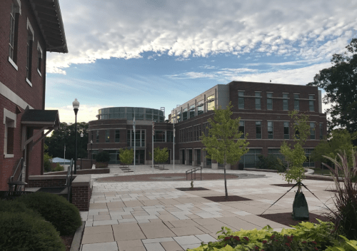 A modern brick building with large windows, surrounded by trees and a paved plaza under a cloudy sky.