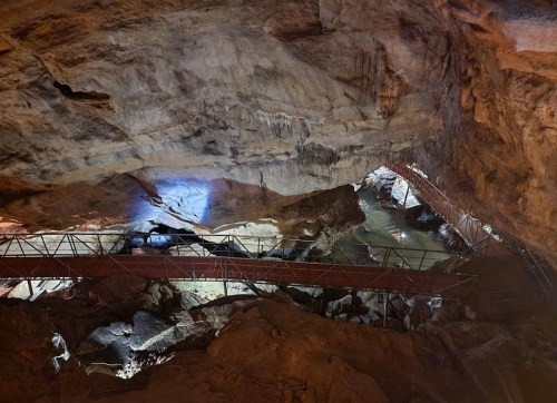 A dimly lit cave interior featuring rock formations, a water body, and a metal walkway.