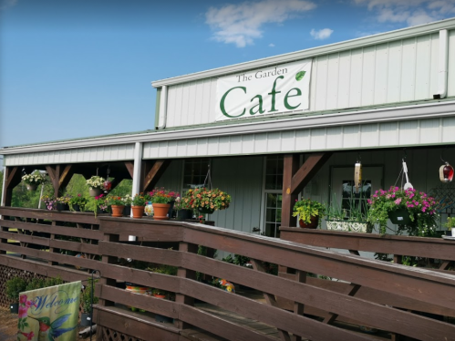 A charming café with a sign reading "The Garden Café," surrounded by colorful flowers and plants on a sunny day.