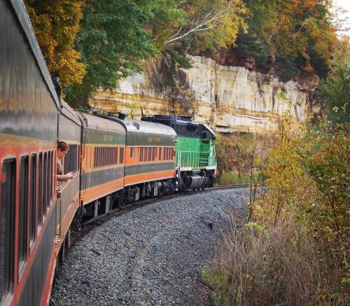 A vintage train curves around a rocky hillside, surrounded by colorful autumn foliage.
