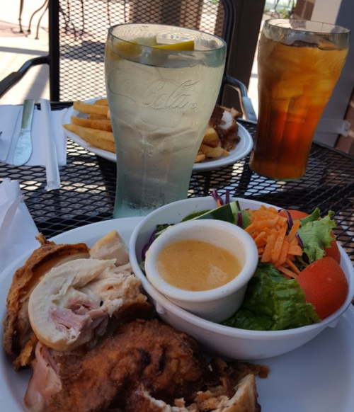 A plate with fried chicken, salad, and dressing, alongside drinks and fries on a table outdoors.