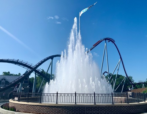 A large water fountain sprays in front of a roller coaster against a clear blue sky.