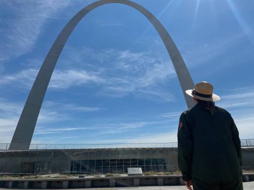A person in a ranger hat stands before the Gateway Arch, gazing up at the towering structure against a blue sky.