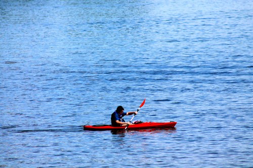 A person paddles a red kayak on a calm blue lake under clear skies.