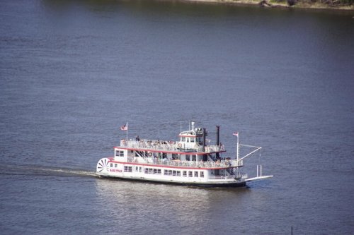 A paddleboat cruises on a river, with passengers enjoying the scenic view on a sunny day.