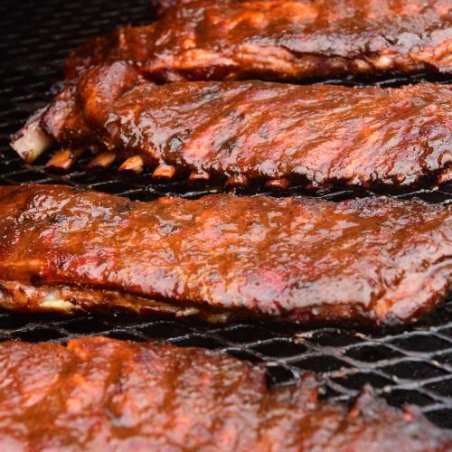 Close-up of glazed barbecue ribs cooking on a grill, showcasing a rich, smoky texture and deep red color.