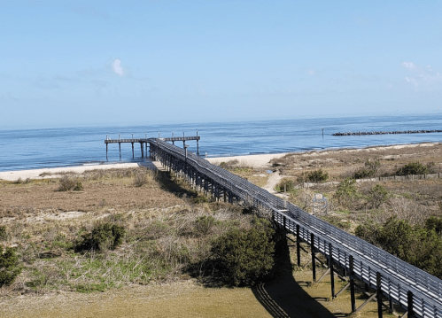 A wooden pier extends over calm waters, leading to a sandy beach under a clear blue sky.