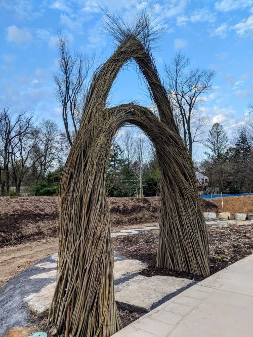 Two large, arching structures made of woven branches, set against a cloudy sky and surrounded by bare trees.