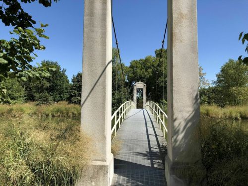 A suspension bridge surrounded by greenery, leading to a stone archway under a clear blue sky.