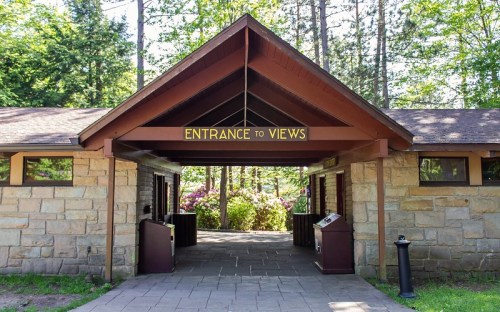 A stone building entrance with a wooden roof, sign reading "ENTRANCE TO VIEWS," surrounded by trees and greenery.