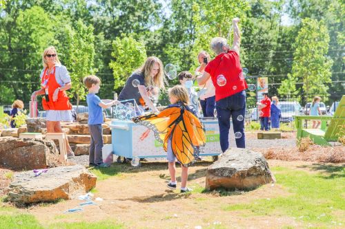 Children and adults play with bubbles in a sunny outdoor setting, with one child wearing a butterfly costume.