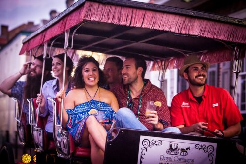 A group of six people enjoying a ride in a festive carriage, smiling and holding drinks in a lively street setting.